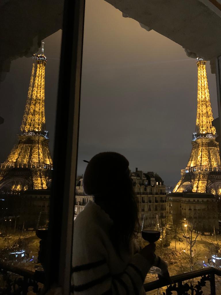 Me and Paname; a window framed and reflecting at night with the Eiffel Tower illuminated, holding a glass of wine wearing French stripes and beret, capturing peak Parisian charm.