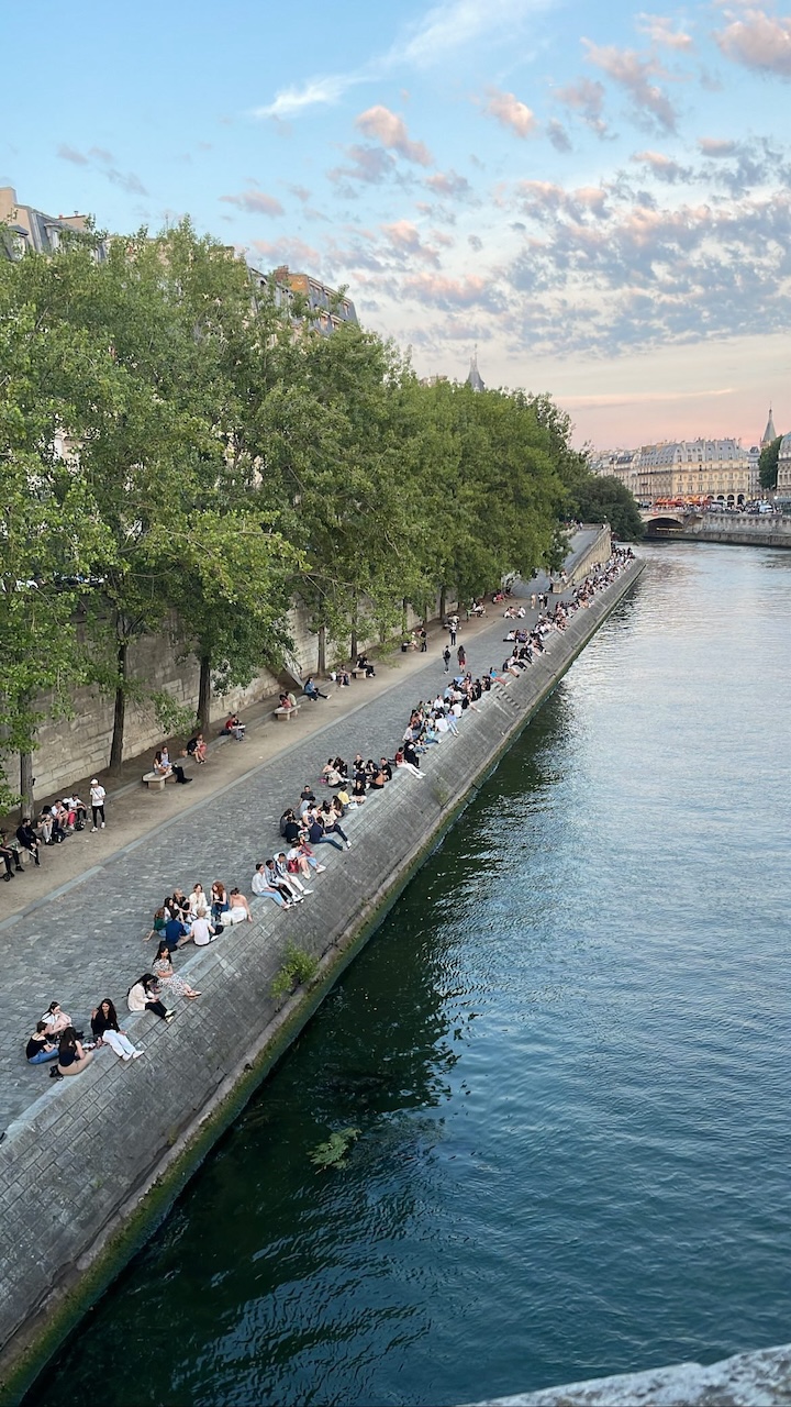 People gathered along the Seine River at sunset during the Summer Solstice in Paris, enjoying music, picnics, and cotton candy skies during Fête de la Musique.