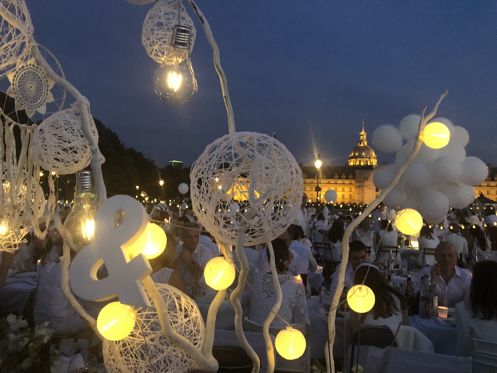 Night falls over Dîner en Blanc Paris 2018 as candlelight flickers across beautifully decorated tables filled with flowers, food, and champagne. The gathering reflects Gemini's connection to communication, social exchange, and the magic that happens when people come together under one sky. Photo by Leah Gillis, The Diamond Is You.