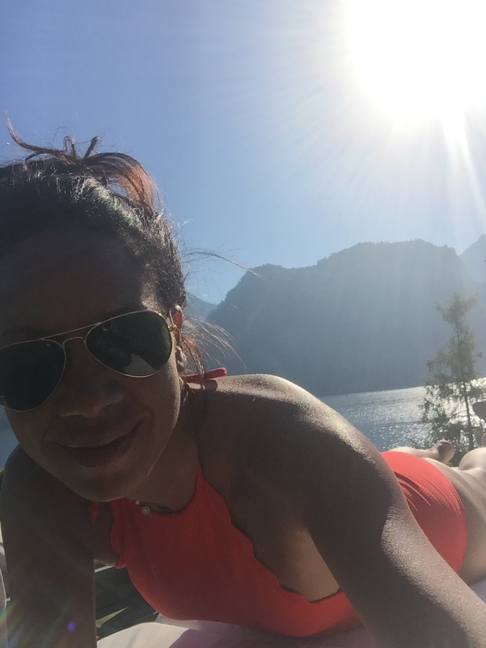Leah Gillis standing above a deep blue alpine lake in Austria wearing an orange bathing suit and sunglasses, with bright sun rays over the surrounding mountains