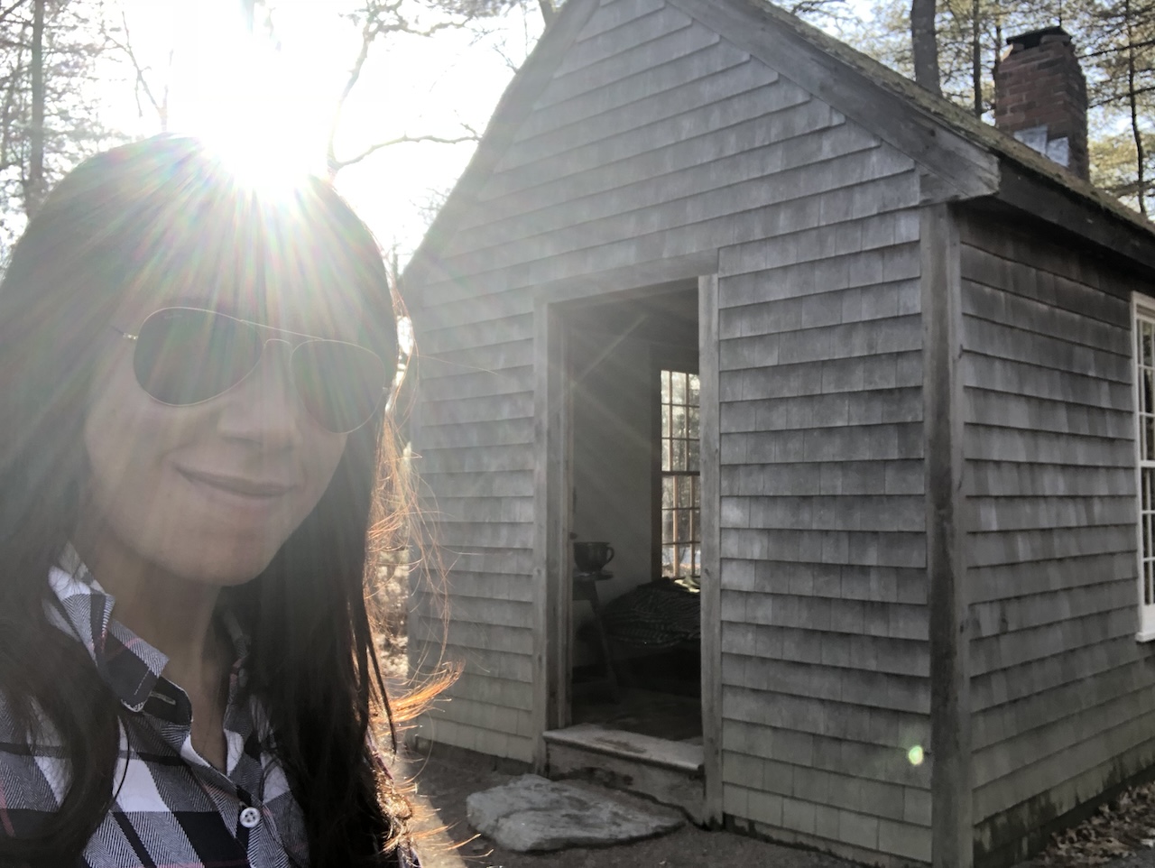 Leah Gillis standing beside Henry David Thoreau's house at Walden Pond as sunlight streams through the trees during Leo season, symbolizing personal leadership, purpose, and harmony with nature.